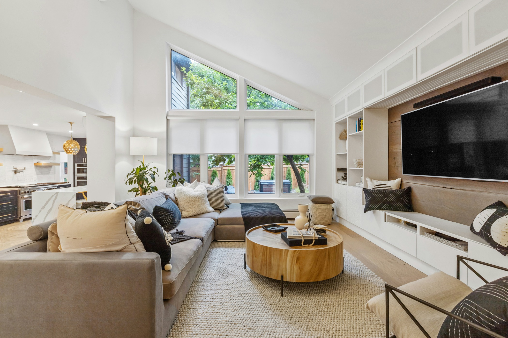 A bright, high-ceilinged living room with a sectional sofa, round wood coffee table, and large windows