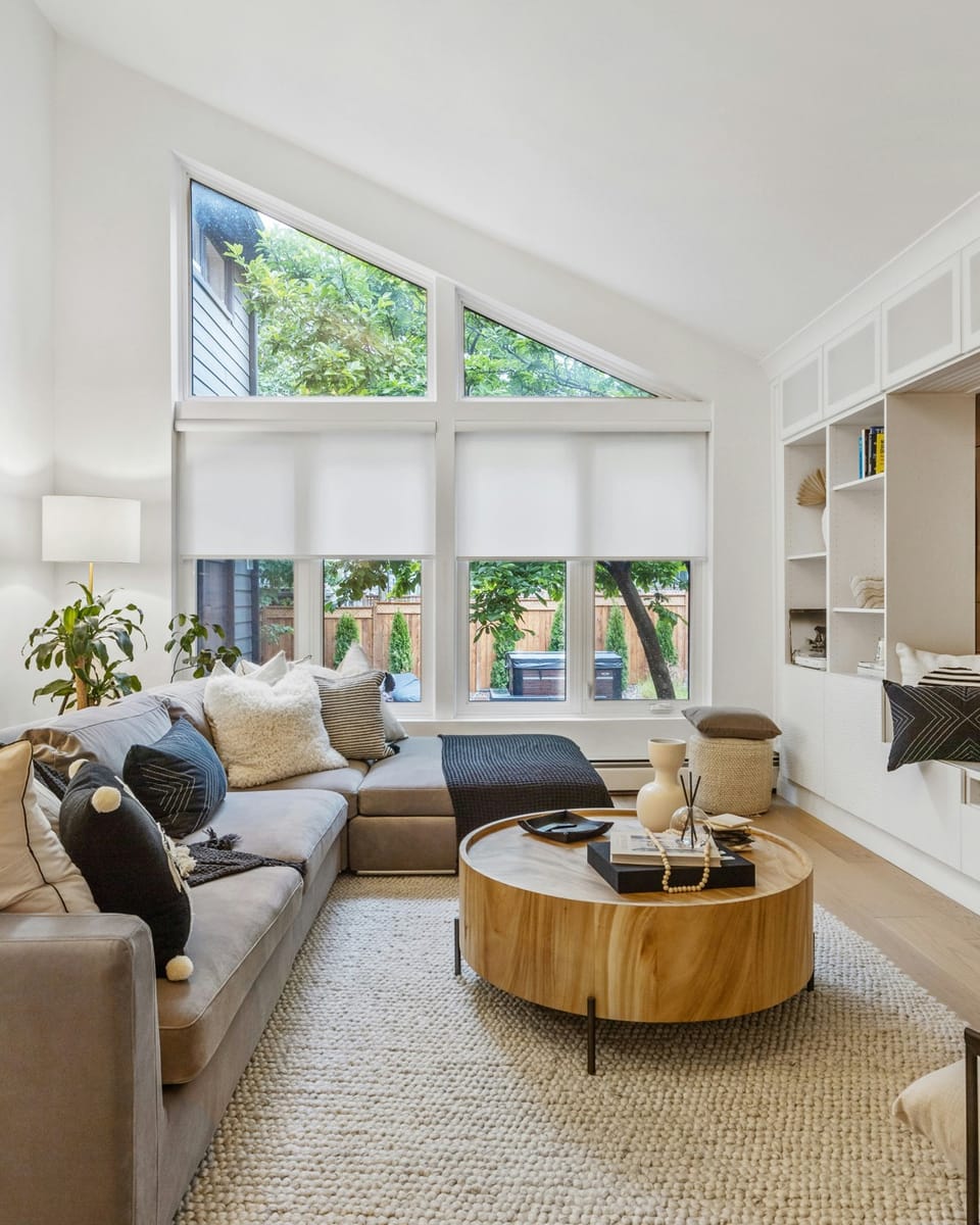 A bright, high-ceilinged living room with a sectional sofa, round wood coffee table, and large windows